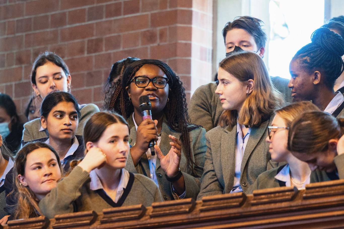 A CLC Sixth Form student with a microphone asks a question at a debate, surrounded by a group of peers