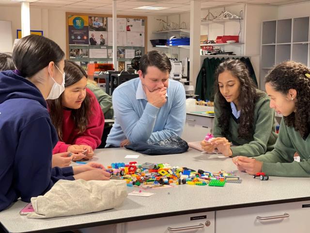 Four students and their teacher sit around a table with Lego pieces on, holding pieces and talking